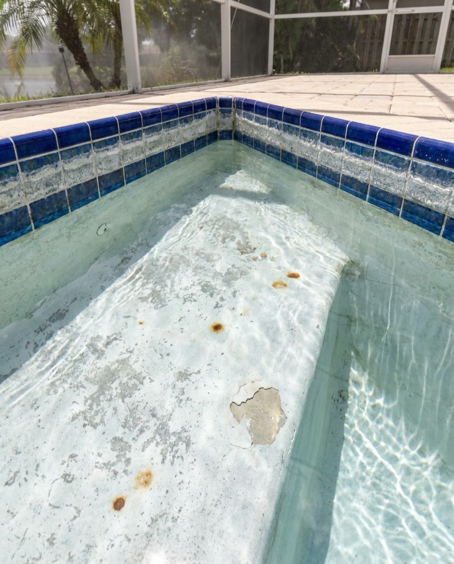 Worn white plaster pool floor, rough and rust-spotted with a flaking patch, under clear shallow water